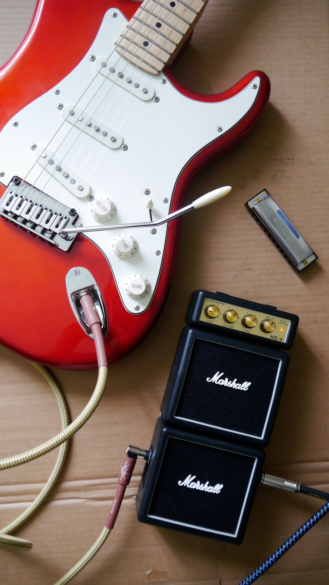 Red electric guitar with cables, a small amplifier and a harmonica on cardboard